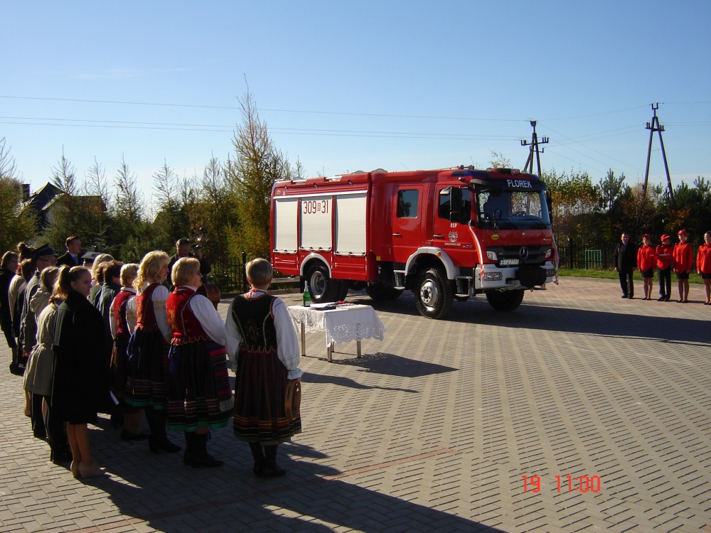 Na zdjęciu widzimy wóz strażacki oraz ludzi. Jest to oficjalne przekazanie nowego wozu strażackiego.