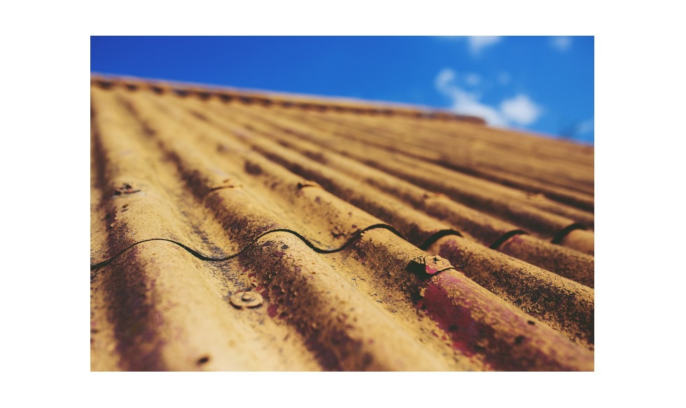 Zdjęcie bliskiego planu dachu falistego z azbestu, dach jest koloru rdzawego i widoczne są kilka śrub i gwoździ. Zdjęcie jest zrobione z niskiego kąta, patrząc w górę na dach. Tło to niebieskie niebo z kilkoma chmurami.