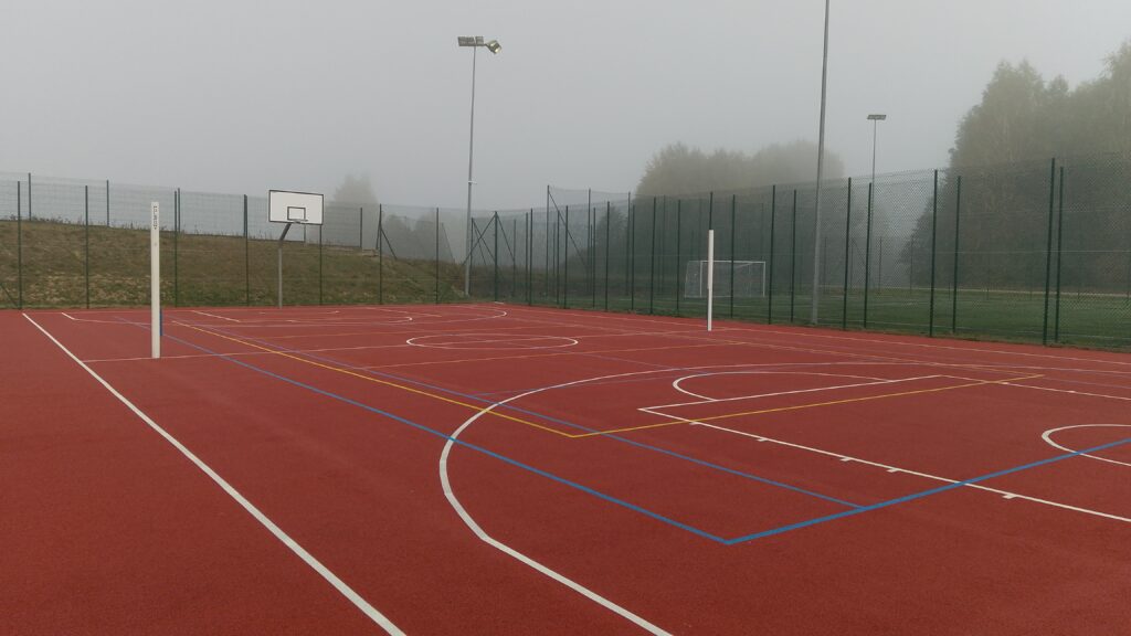 Wielofunkcyjne boisko sportowe na świeżym powietrzu z oznaczeniami do koszykówki, tenisa i piłki nożnej, otoczone ogrodzeniem i zielenią w lekko zamglonym krajobrazie.