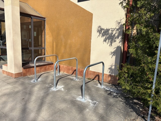 Wejście do budynku z przeszklonym przedsionkiem po lewej stronie. Obok drzwi na chodniku znajdują się 3 stojakami na rowery typu