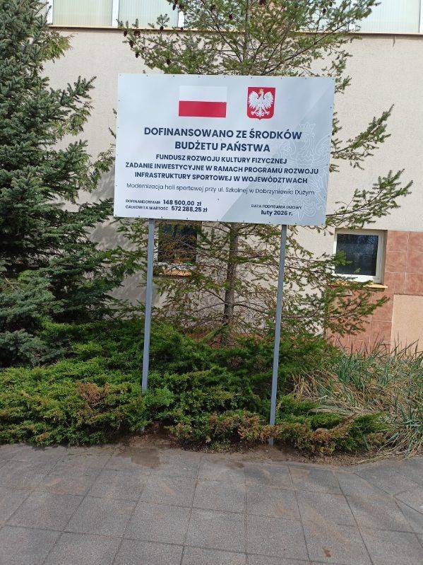 Tablica informacyjna z logo Polski i godłem państwowym, informująca o dofinansowaniu inwestycji sportowej za środków budżetu państwa. Znak stoi na dwóch metalowych słupach przed budynkiem, w otoczeniu zieleni- krzewów i małego drzewa