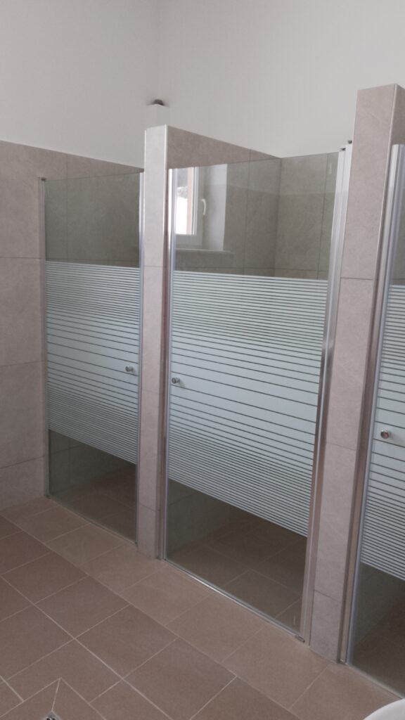 3 Kabiny prysznicowe w łazience, odgrodzone ściankami z jasnymi płytkami na ścianach i podłodze.