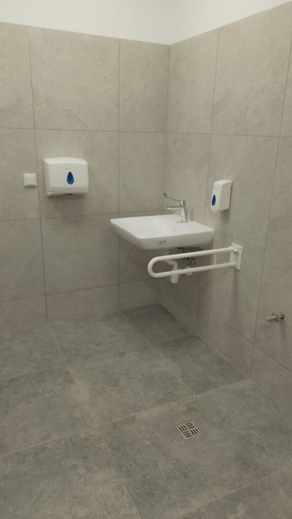 Umywalka w łazience przystosowananej dla osób z niepełnosprwwnościami.  Łazienka z jasnoszarymi płytkami na ścianach i podłodze.
