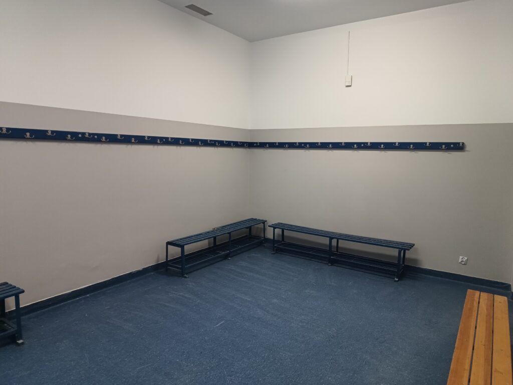 Pusta szatnia z jasnoszarymi ścianami, brązowymi wieszakami na ubrania i metalowymi ławkami,
ustawionymi przy ścianach z pomarańczową podłogą. 