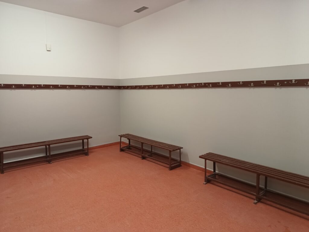 Pusta szatnia z jasnoszarymi ścianami, niebieskimi wieszakami na ubrania i metalowymi ławkami,
ustawionymi przy ścianach z pomarańczową podłogą.