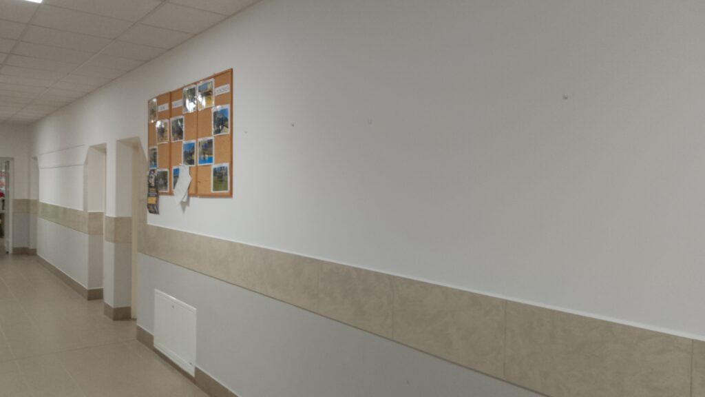 Korytarz na którym znajduje się tablica ogłoszeń z pracami uczniów.
Dominują jasne kolory ścian i podłogi, z beżowym pasem glazury i lamperii w dolnej części ścian. 