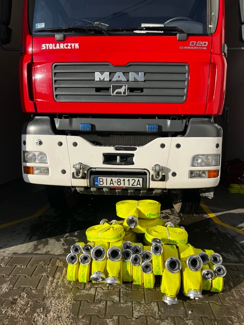 Na zdjęciu widać przód czerwonego samochodu pożarniczego przed którym ułożone są w kilku rzędach żółte węże pożarnicze z nasadami